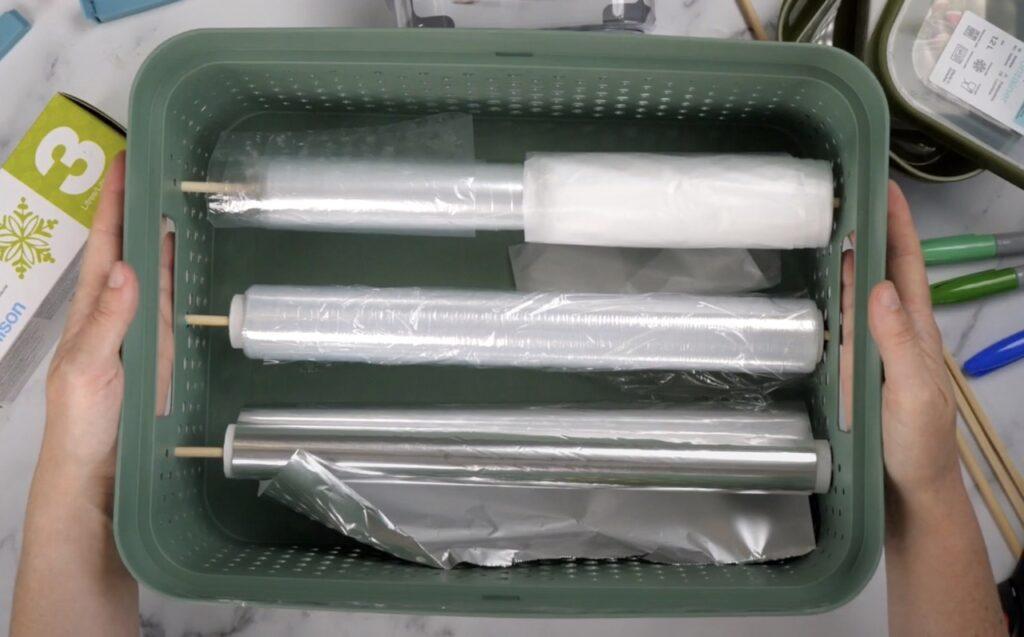 Bilde med plasposer, plastfolie og alluminiumsfolie i en gønn kurv. Vi viser deg kreative måter å organisere kjøkkenet på.