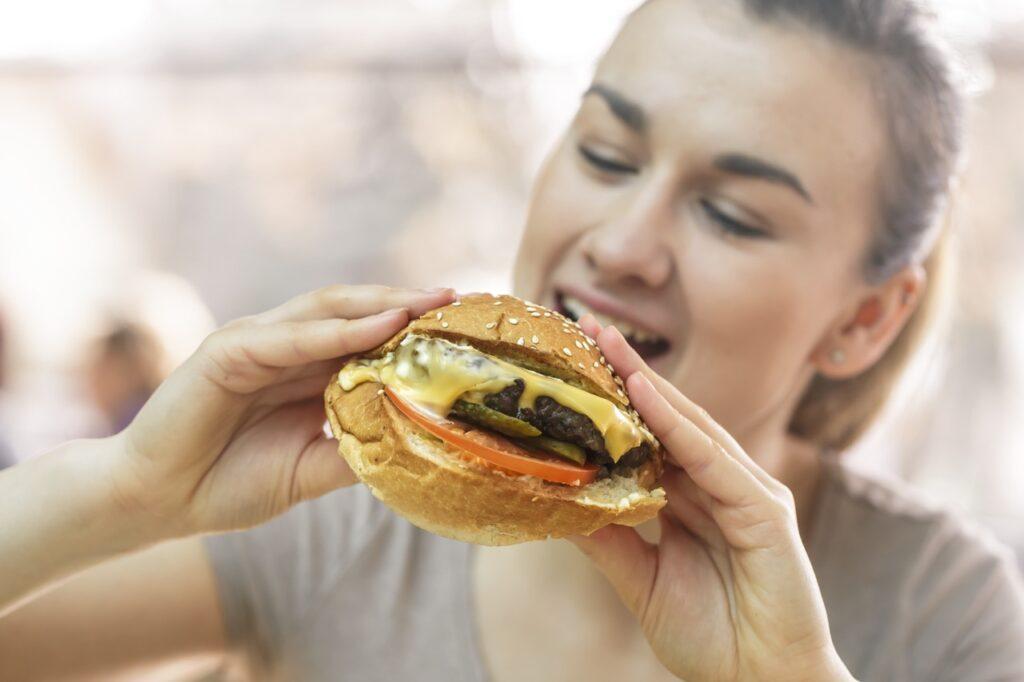 Bilde av blid kvinne som skal til å ta seg en bit av en hamburger med tomat, smeltet ost og sylteagurk til artikkelen som forklarer hvorfor noen mennesker aldri legger på seg.