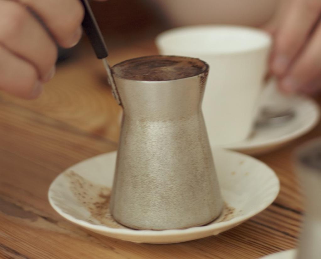 Bilde av verdens sunneste kaffe, gresk kaffe. En hand holder i håndtaket til en liten kanne med gresk kaffe på vei til å helle den i en hvit kopp på et bord.