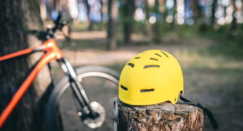 Bilde av gul sykkelhjelm på stubbe foran elsykkel som er lent mor et tre ute i naturen. Vi viset hvordan du velger riktig hjelm til elsykkel.