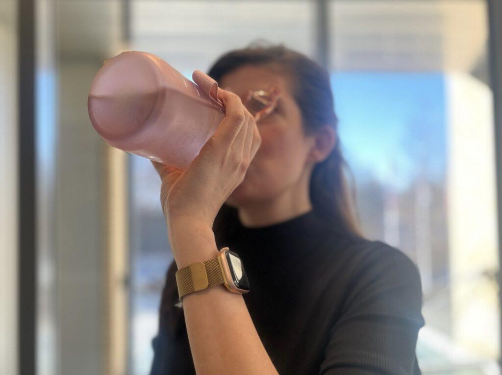 Bilde av en dame som drikker vann av en rosa motivasjonsflaske. Hun har på seg en svart genser og en apple watch i gull, og står foran et vindu. Vi forteller om «vanntabben» som kan send deg opp i vekt! Og forteller hvordan det å drikke vann riktig eller feil kan ha innvirkning på forbrenningen din. 