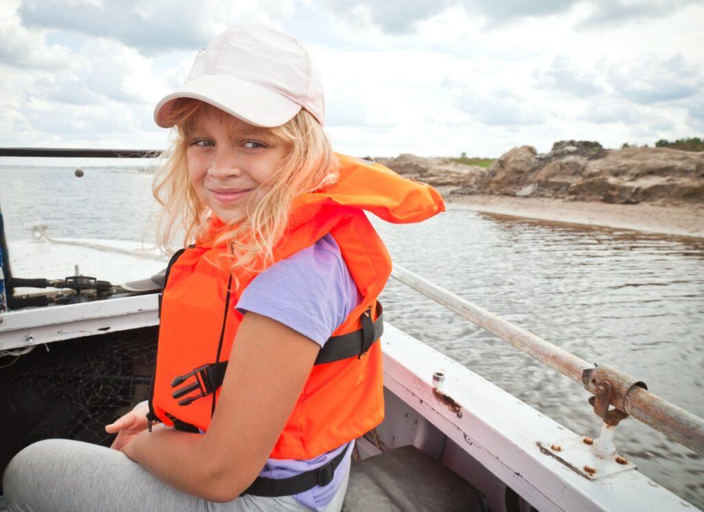 Smilende blond jente med hvit caps og orange redningsvest ombord i en liten båt. Vi hjelper deg med å finne riktig flytevest eller redningsvest slik at sommeren på sjøen blir tryggest mulig.