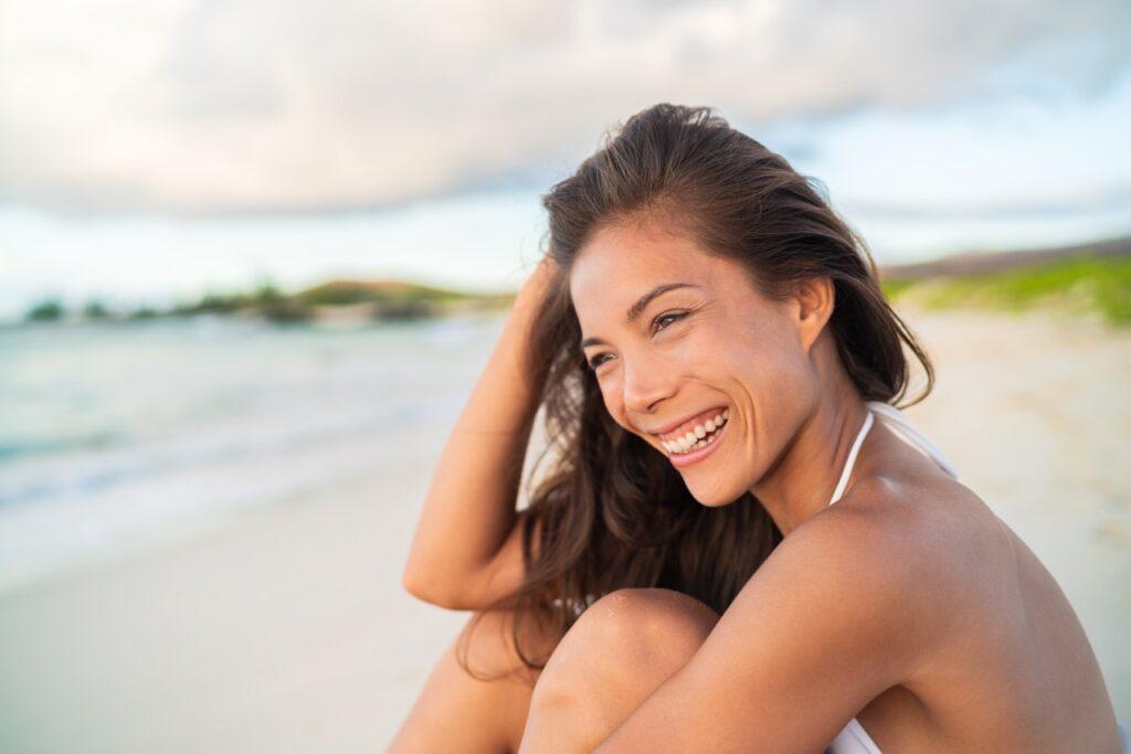 Bilde av kvinne smilende på en sandstrand, solbrun om sommeren. Med sandstrand og hav i bakgrunnen.