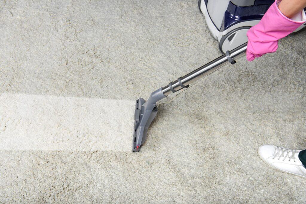 Bilde av en hvit skittent gulvteppe hvor en person med rosa hansker bruker en tepperenser for å rense teppet. På teppet ser du tydelig en ren stripe hvor hun har brukt damprenseren. Vi har samlet de beste tepperenserne ifølge eksperter og forbrukeren.