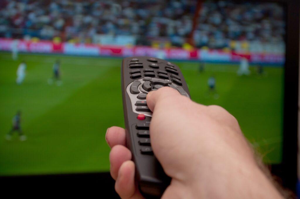 Viktige begreper i fotball: Hånd med fjernkontroll peker mot en TV-skjerm som viser fotball.