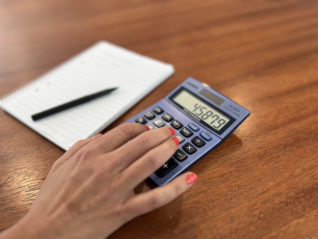 Bilde av en hand med rød neglelakk som regner sammen tall på kalkulator for å finne ut om man betaler for mye i renter på lån. Vi viser deg hvordan du kan forhandle om bedre rente på lånene dine for å spare penger ved rentehevingen i 2023.