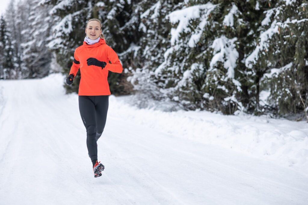 Bilde av lyshåret dame som løper ute om vinteren. Hun løper på snø mellom snødekte grantrær og smiler i en rød jakke og svarte bukser. Vi avslører hvilke joggesko og løpesko med pigger som er best i test i 2023. Vinnerne er fra Saucony, La Sportiva, Nike og Inov-8.