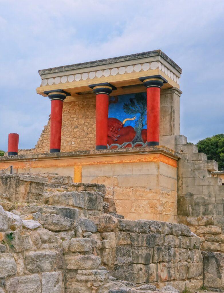 Bilde av ruiner i Knossos på Kreta i Hellas. Fargene på ruinene er enda tydelige med røde søyler og blå fresker. Vi har funnet restplasser til Kreta slik at du kan nyte billig reise til Hellas. Vi gir deg også fem tips til ting du må se og gjøre på den vakre øya, blant annet: Knossos, Samariaravinen, Elafonissi, Rethymnon og Spinalonga.