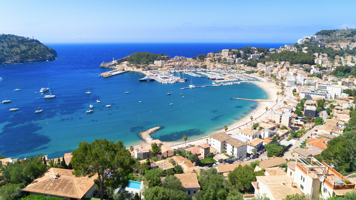 Bilde av en liten middelhavsby på den spanske øya Mallorca. På bilde ser du knallblått hav, en hvit strand. båter på havet og en koselig liten by som strekker seg opp i fjellet.