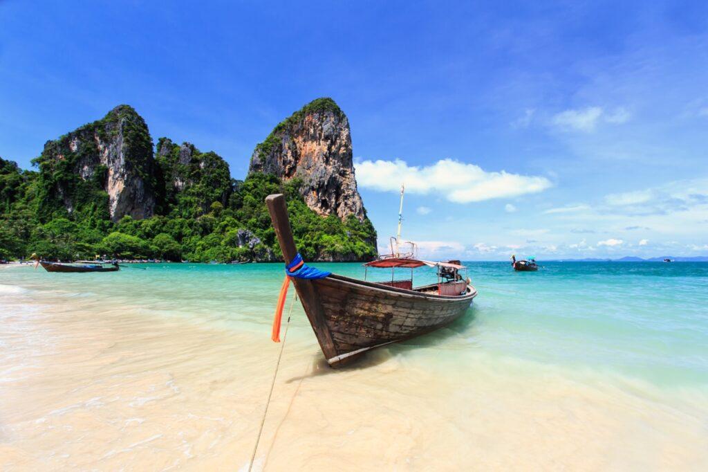 En klassisk thailandsk båt som ligger ved en hvit strand med knallblått hav og klipper i bakgrunnen. Vi har funnet restplasser til Thailand slik at du kan finne billige pakkereiser til den populære feriedestinasjonen.