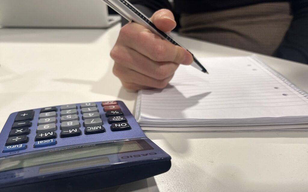 Bilde av en person i svart genser som sitter ved en pult og regner på gjeld med en notatblokk, en penn og kalkulator. Vi forteller hva du må vite om din egen gjeldsgrad og hvor enkelt du sjekker det gratis. 