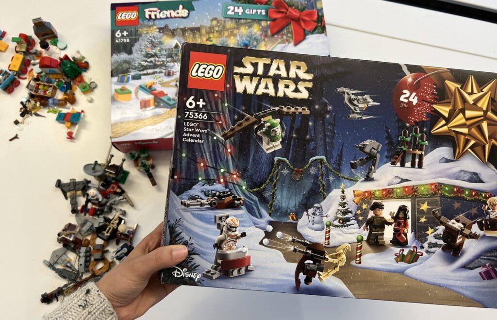 Hånd som holder en LEGO Star Wars julekalender. I bakgrunnen ligger en julekalender med LEGO Friends samt flere LEGO-deler ved siden av. Vi har åpnet julekalendere fra LEGO for å finne ut om de er verdt pengene og holder det som blir lovet. Vi ville finne ut om barna blir glad for de populære kalenderne!