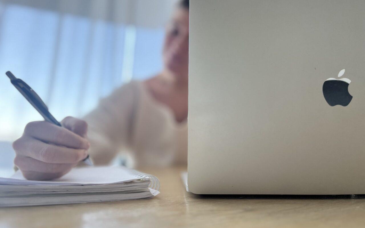 Bilde av kvinne som sitter med en Macbook ved et bord og regner på en notatbok. Vi forteller når og hvorfor det kan lønne seg å refinansiere smålån ved hjelp av å samle alle til ett større lån.
