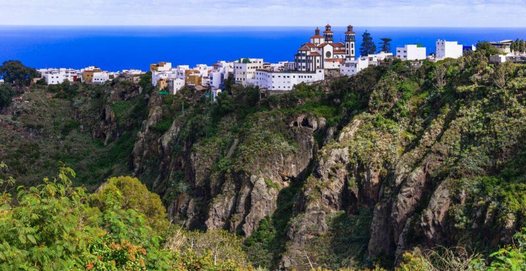 Bilde av landsby i frodige åser på Gran Canaria. Her ser du majestetiske fjell, grønne trær og busker og rosa blomster. Vi ahr funnet frem forslag til billige pakkereiser som du får billig med restplasser til den populære Kanariøyen med besøksfavoritter som Puerto Rico, Las Palmas og Playa del Inglés.