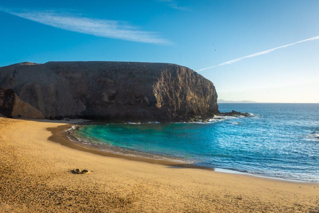 Bilde av en strand med turkist hav, i bakgrunnen kan man se et berg av lavastein. Vi har snakket med redaktør i Lanzaroteposten, Ola Kjeldstad som gir oss sine beste tips til en topp 5 Lanzarote-guide.