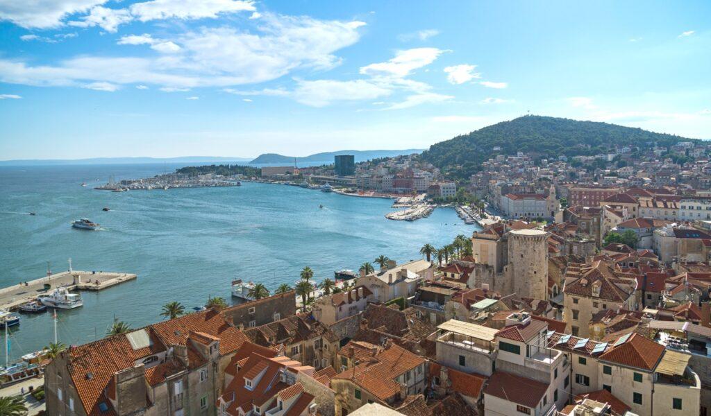 Utsiktsbilde over Split i Kroatia fra Marjan-høyden. Her ser du endeløst blått hav. Bygninger med røde tak og en grønn ås i bakgrunnen. Split tilbyr alt fra lokale markeder, strender, utsiktspunkt, gamle romerske ruiner og en kort båt tur til en av verdens kanskje fineste strender. Vi gir deg 5 tips til ting du må se og oppleve i byen, tips til hvor du finner billige reiser, samt svar på ofte stilte spørsmål om Split.