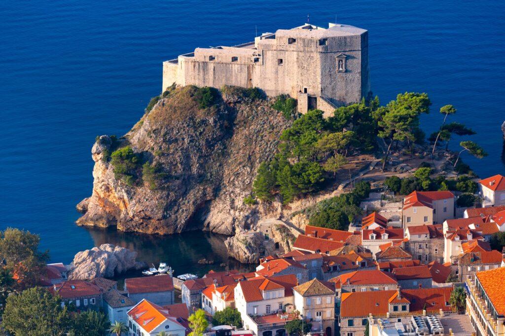 Bilde av Fort Lovrijenac (Dubrovnik, Kroatia) i bakgrunnen som står på en klippe med blått hav rundt. I forgrunnen ser man klassiske murhus med rødt tak. Dubrovnik byr på alt fra vakre strender til historiske severdigheter som Fort Lovrijenac, som også har blitt brukt som sett i den populære serien Game of Thrones.