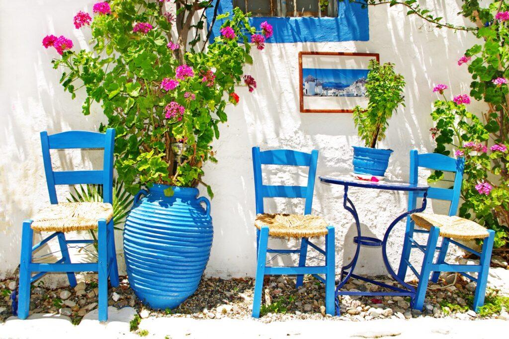 Bilde av tre blå stoler som står langs en hvit murvegg på Kreta i Hellas. På murveggen kan man se et vindu med blå vinduskarm, samt en stor blå krukke med grønn plante med rosa blomster. Vi har laget en lang liste på hvor du må reise for å finne varme. Vi avslører blant annet temperaturer på Gran Canaria, Hellas, Kroatia og mange mange flere populære reisedestinasjoner.