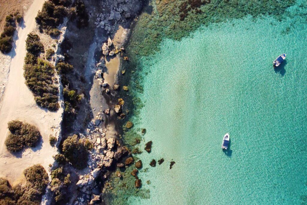 Luftbilde av Akamas-halvøya på Kypros, som viser en imponerende kystlinje med krystallklart turkist hav og to hvite båter som flyter nær kysten, noe som fremhever den rolige skjønnheten ved dette populære reisemålet. Vi hjelper deg med å finne restplasser til Kypros slik at du kan finne billige reiser til syden.