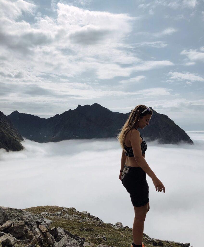 Bilde av Runa Sommerfelt Ovenstad som står på toppen av et fjell med utsikt over skyer og andre fjell. Runa har gitt oss sine beste tips til hva du må ha på pakkelista om du planlegger fjellturer i sommer. Hun avslører også sine favoritt-fjellturer i Norge!