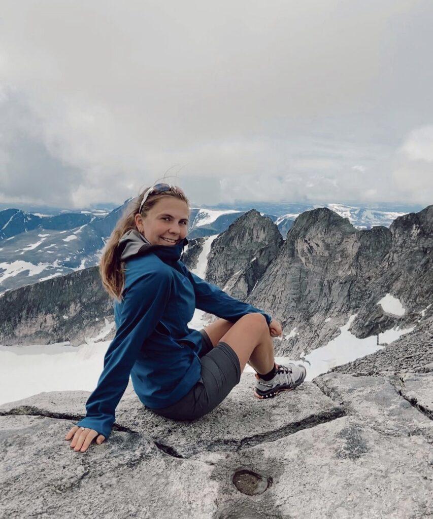 Bilde av Runa Sommerfelt Ovenstad på toppen av et fjell på Offersøykammen i Lofoten. Smilende mot kamera i en rosa te-skjorte og med en vakker solnedgang i bakgrunnen. Runa forteller hva du må ha på pakkelisten når du skal på fjelltur i sommer i tillegg til å dele sine beste tips og favoritt-fjellturer i Norge.