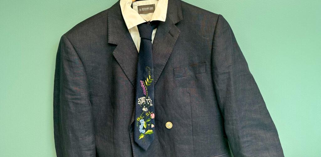 Mørk dressjakke, hvit skjorte på en kleshenger med et mørkeblått slips med broderte bunadsblomster på.