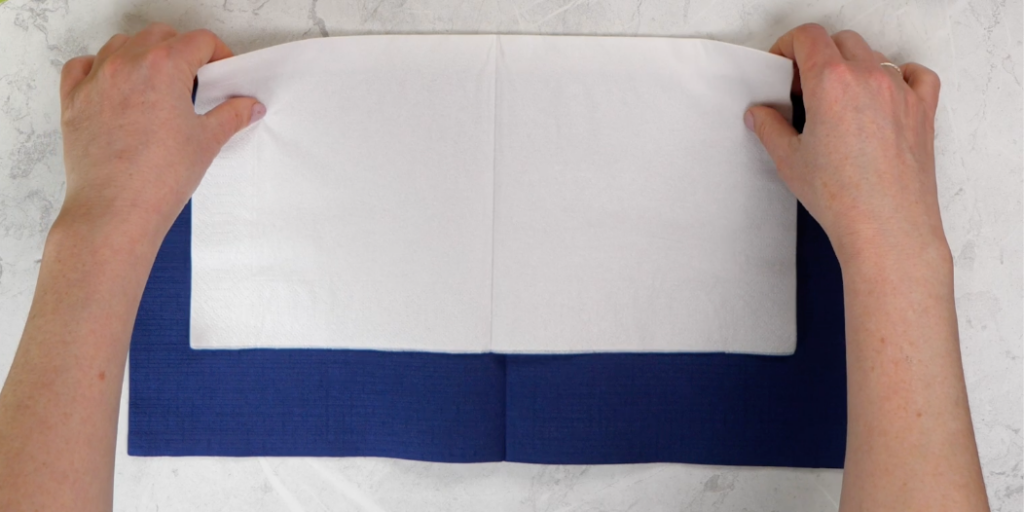 To servietter i ulik farge og størrelse, blå og hvit, som ligger oppå hverandre. Hender i ferd med å begynne å brette serviettene til en bunad.