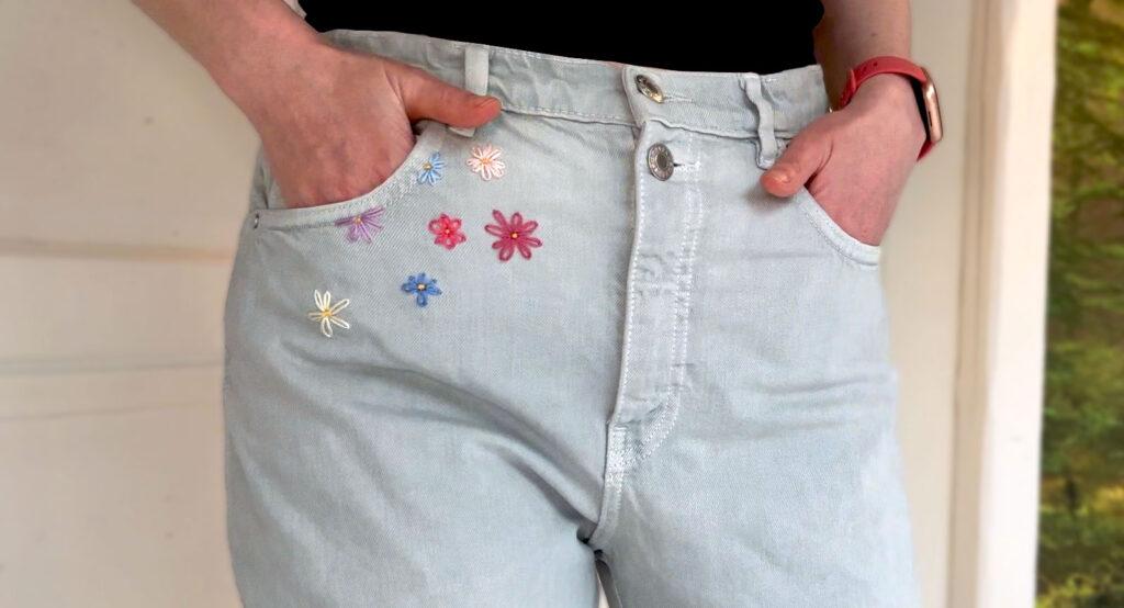 Person med lyse jeans, som har hendene i lomma. Ved bukselommen er det bordert blomster i ulike farger.