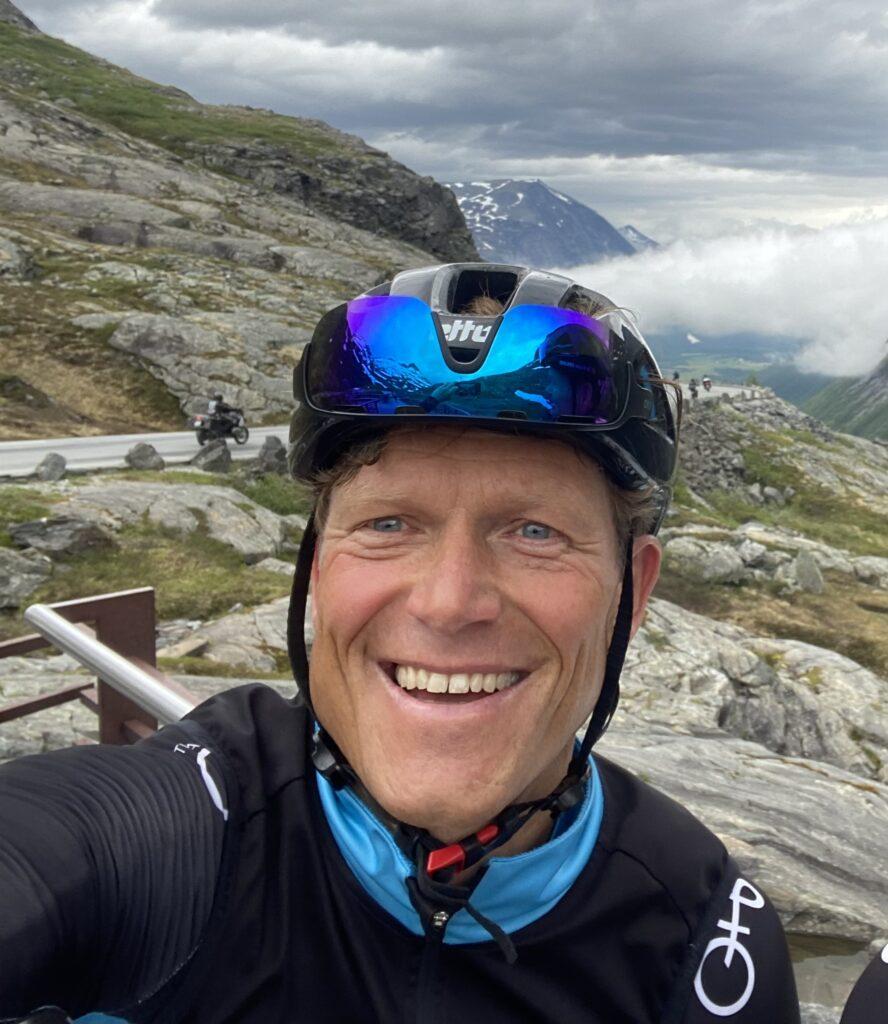 Bilde av Ola G. Bustad smilende med sykkelhjelm på toppen av hodet op toppen av Trollstigen. Vi spurte Bustad hva man må tenke på når man velger sykkel til barn, hvordan man vleger riktig størrelse eller hvor mange år og hvilken alder barnet har. Han forteller også litt om både vekt og dekk på sykkel!