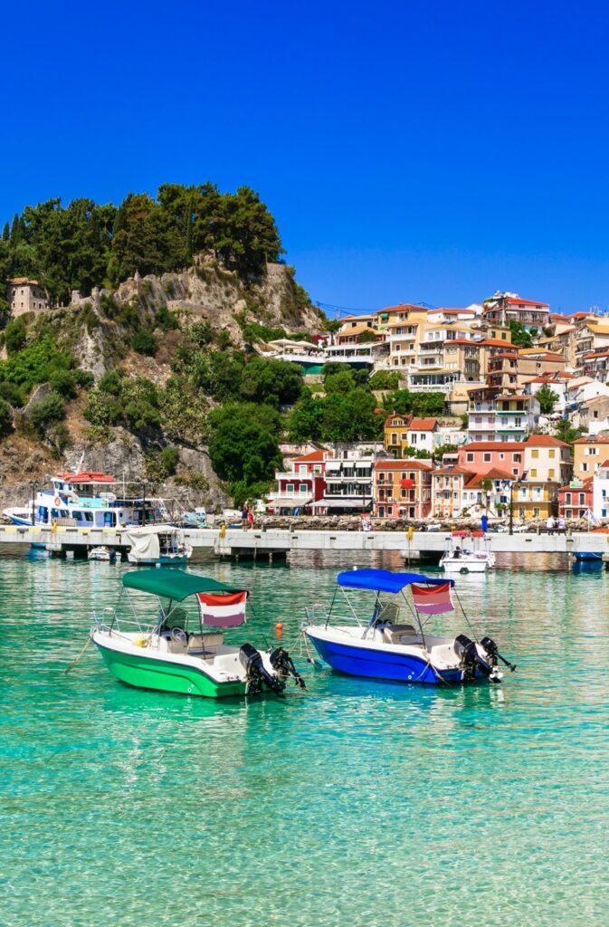 Bilde tatt fra den krystallklare sjøen ved Parga i Hellas. I forgrunnen ser du en grønn og en blå båt, og i bakgrunnen kan du se fargerike murhus i byen, samt grønnkledde åser. Vi gir deg fem ting vi mener du bør se og gjøre i Parga, blant annet Parga-borgen, Ali Pasha-borgen og besøke de mange strendene. Vi gir deg også tips til hvor du finner pakkereiser og restplass-reiser.