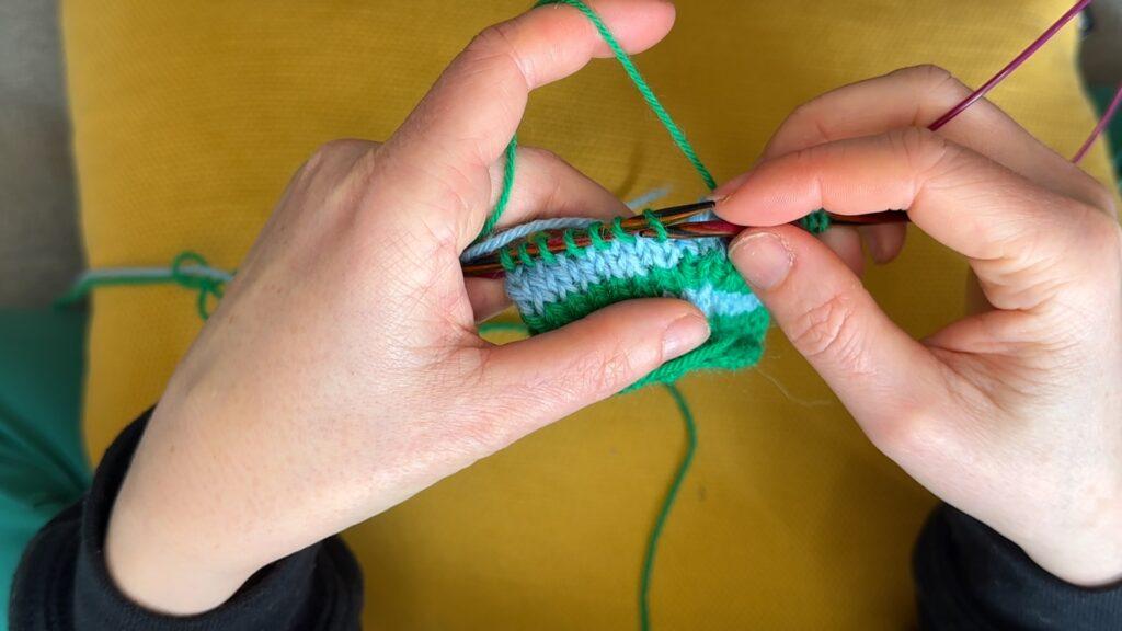 Hender som holder strikketøy med striper i blått og grønt.