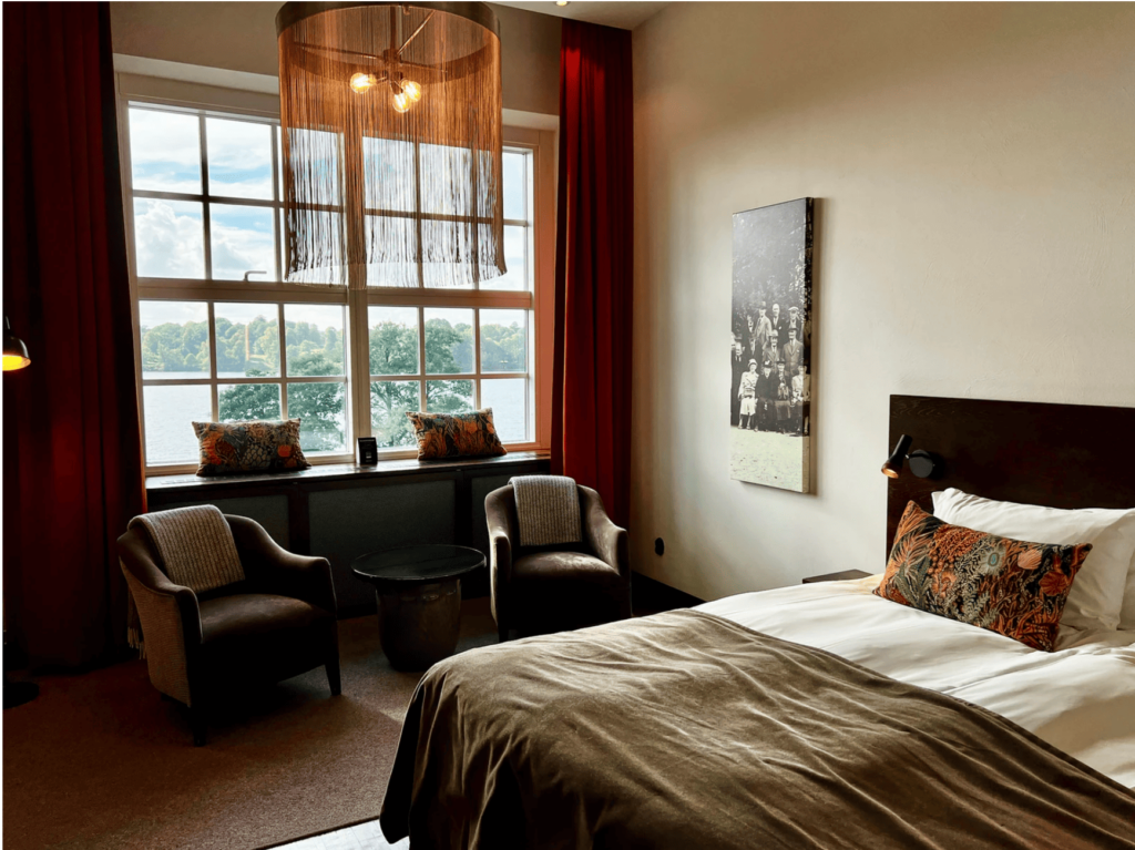 Et komfortabelt hotellrom med utsikt over innsjøen, dekorert med stilrene detaljer og en stor vindusflate.