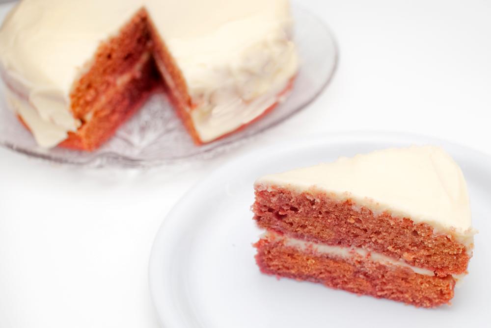 Vi deler oppskrift på vegansk red velvet kake. Her er et stykke av kaken satt frem på en tallerken, men resten av kaken står i bakgrunn.